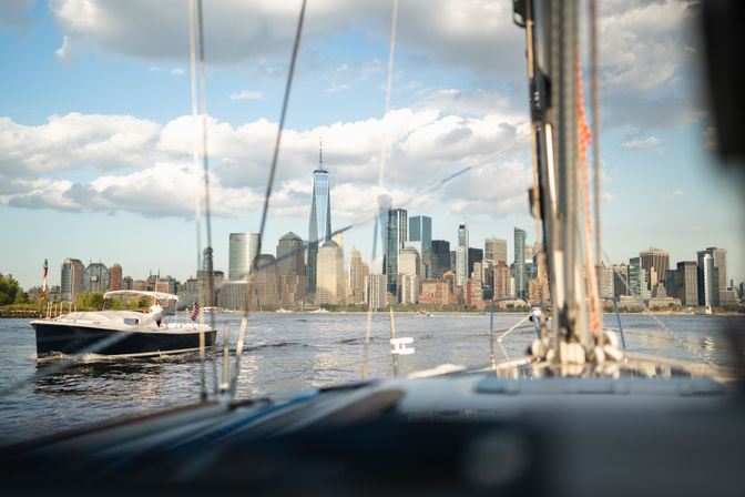 Sailboat view toward Manhattan skyline and One World Trade Center across the Hudson River with a nearby motorboat and sunny, cloud-streaked sky