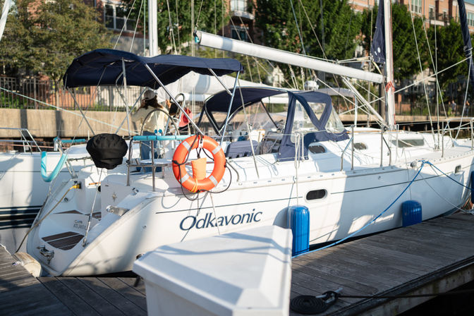 White sailboat docked at a sunny marina with navy canvas covers, orange lifebuoy, blue fenders and a wooden dock