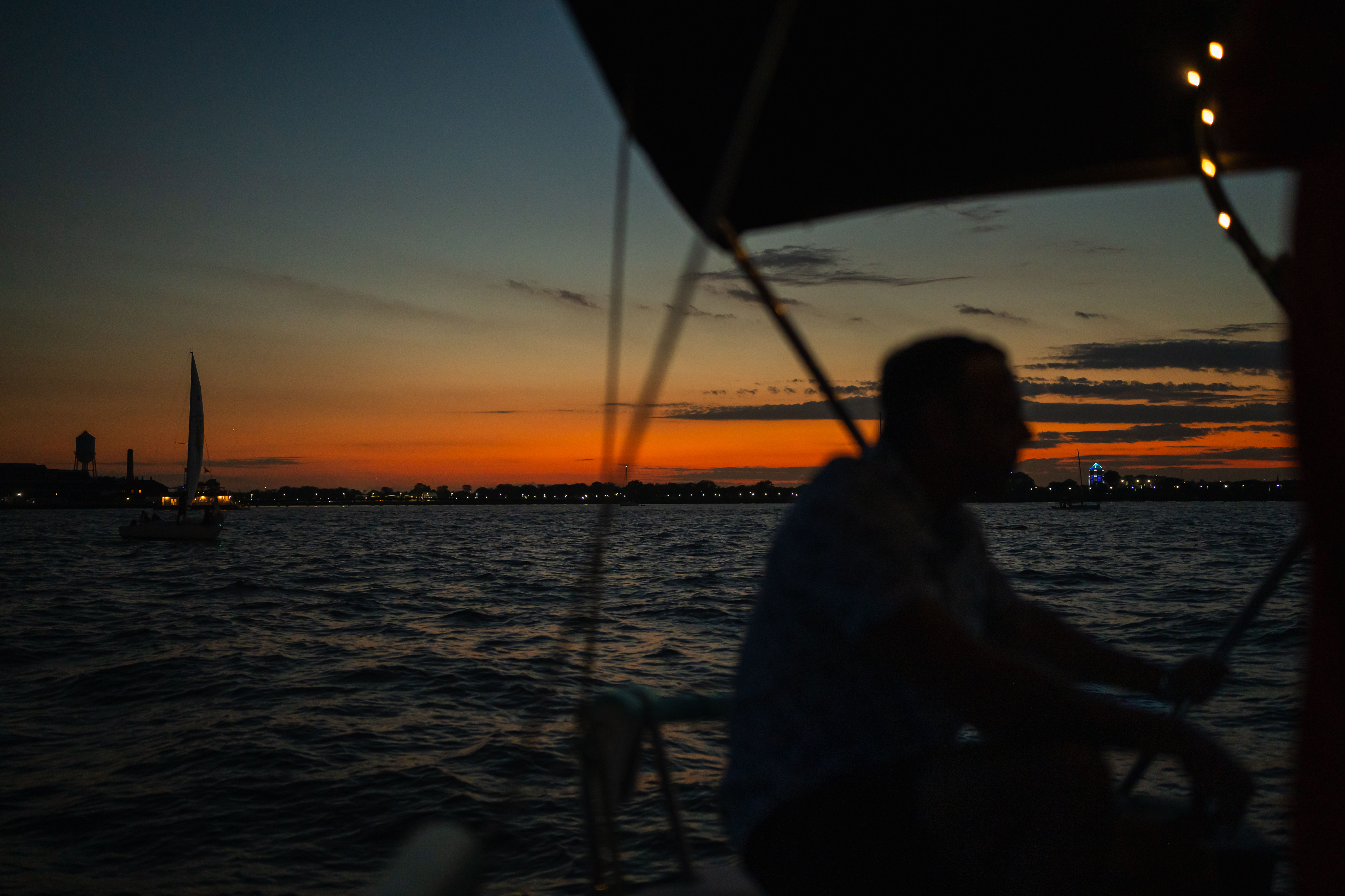 Silhouetted person on a sailboat at sunset, orange-and-blue sky over choppy water with distant shoreline lights and another sailboat on the horizon