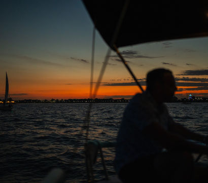 Silhouetted person on a sailboat at sunset, orange-and-blue sky over choppy water with distant shoreline lights and another sailboat on the horizon