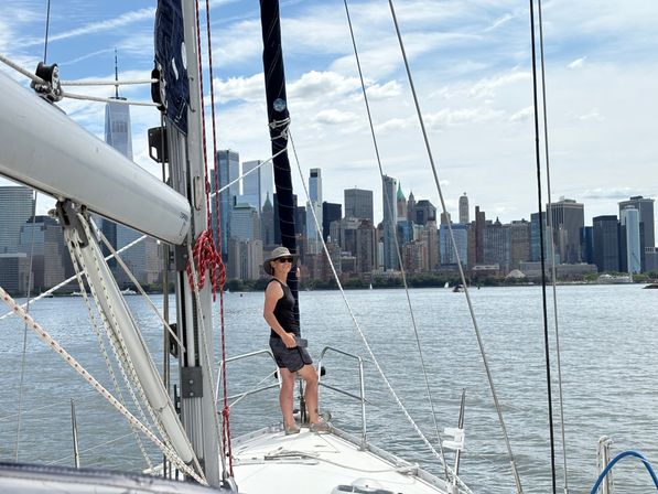 Person in a sunhat standing on the bow of a sailboat on the Hudson River with the Manhattan skyline and One World Trade Center in the background under a bright, partly cloudy sky.