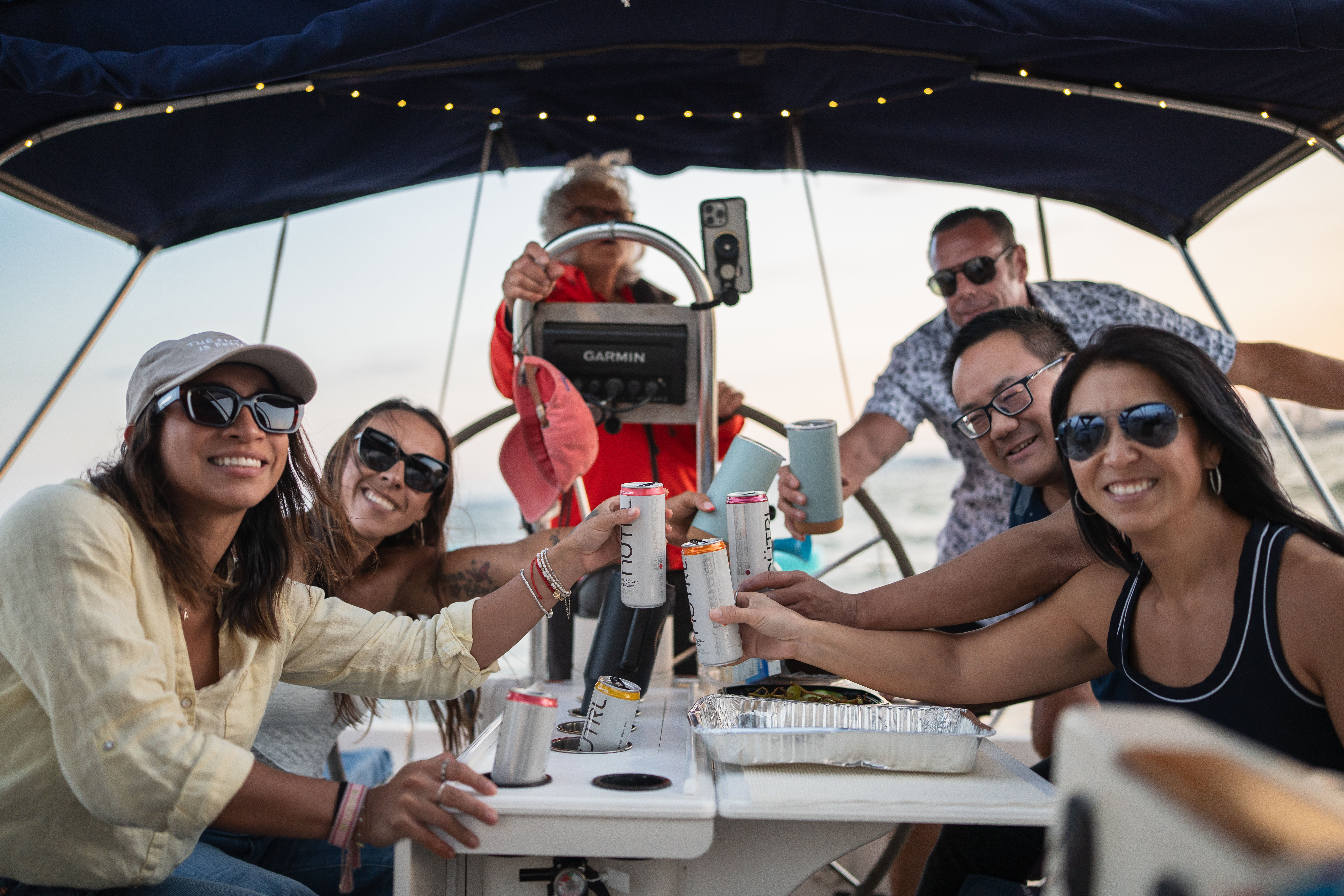 Group of friends on a sailboat at sunset, smiling and toasting with canned drinks in the cockpit near open water