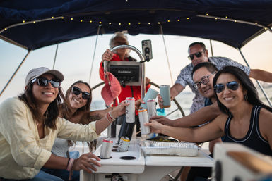 Group of friends on a sailboat at sunset, smiling and toasting with canned drinks in the cockpit near open water