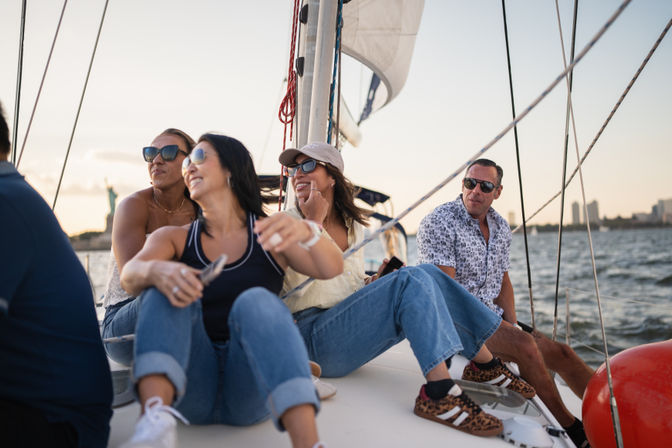 Four adults laughing and relaxing on a sailboat at sunset near the Statue of Liberty with the New York skyline in the background.