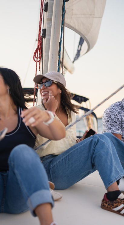 Four adults laughing and relaxing on a sailboat at sunset near the Statue of Liberty with the New York skyline in the background.