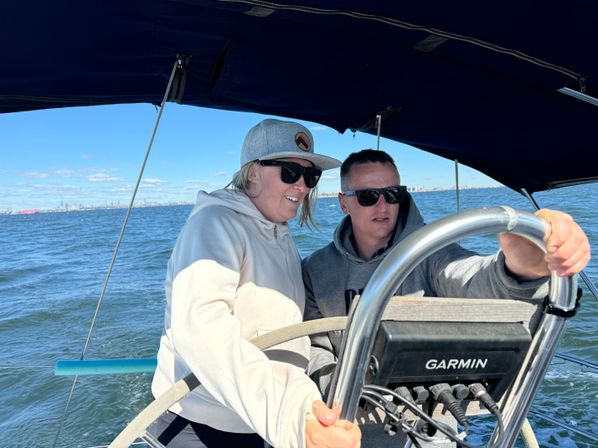Two people in sunglasses steering a sailboat at the helm under a navy bimini on sunny coastal waters with a distant city skyline