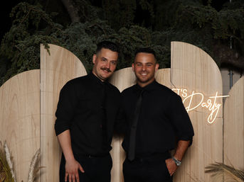 Two smiling men in black shirts and ties pose at an evening outdoor event photo backdrop with curved light-wood panels, greenery overhead, pampas grass accents, and a neon 'Let's Party' sign.