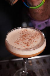 Close-up of a frothy amber craft cocktail in a coupe glass topped with a light cinnamon dusting, sitting on a bar surface.