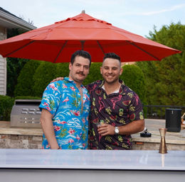 Two friends in colorful Hawaiian shirts smiling at an outdoor backyard patio bar under a red umbrella, one holding a cocktail beside a grill and lush green hedges — summer party vibe.