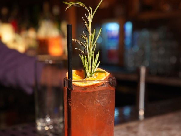 Iced orange cocktail in a tall glass garnished with a rosemary sprig and dried orange slice and a black straw, sitting on a bar counter with warm blurred lights in the background.
