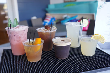 Assorted colorful summer cocktails and mocktails in plastic cups with fruit garnishes (pineapple, orange, lemon, cucumber) served on a black bar mat at a sunny poolside bar