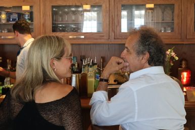 Two patrons chatting at a cozy wooden restaurant bar, bottles, shakers and glassware lined on the counter and shelves behind them.