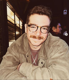 Smiling person with tortoiseshell glasses and a thick mustache, wearing a tan jacket and layered necklaces, leaning on a table in a cozy dimly lit bar.