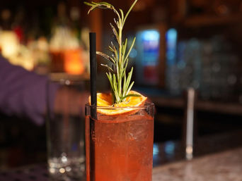Zesty citrus craft cocktail in a highball glass with ice, dried orange slice and rosemary sprig garnish and a black straw on a dimly lit bar counter
