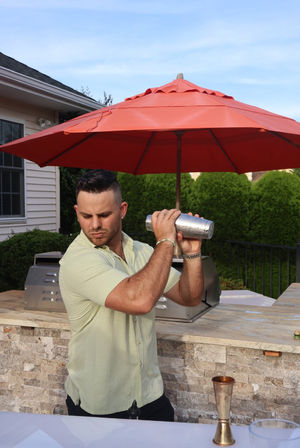 Person shaking a cocktail shaker at a backyard patio bar with a red umbrella, stone countertop and outdoor grill