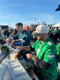 Two fans in green Philadelphia Eagles jerseys pouring drinks at an outdoor tailgate bar, crowd and stadium structures in the background under a clear blue sky