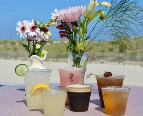 Beachside table with six refreshing colorful summer drinks in clear cups and two small vases of pink and white flowers, sandy dunes and bright blue sky in the background.