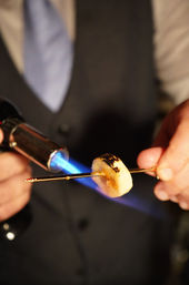 Close-up of hands using a blue culinary blowtorch to caramelize a banana slice on a metal skewer, creating golden, slightly charred spots for dessert prep