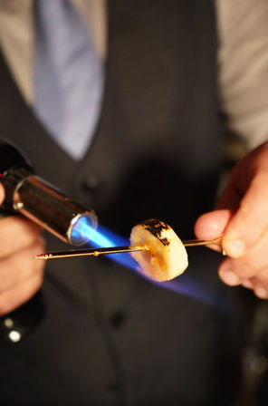 Close-up of hands using a blue culinary blowtorch to caramelize a banana slice on a metal skewer, creating golden, slightly charred spots for dessert prep