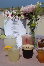Beachside cocktail display with a printed cocktail menu, vase of pink flowers, and several iced mixed drinks in clear cups on a pink tablecloth with sandy dunes in the background.
