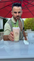 Man on an outdoor patio serving a pastel pink-and-cream tropical cocktail garnished with pineapple leaves beneath a red patio umbrella at a backyard bar