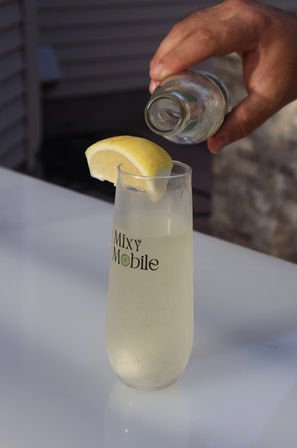 Hand pouring from a small bottle into a chilled champagne flute with a pale lemon cocktail garnished with a lemon wedge on a white outdoor table.