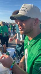 Close-up of a man in a green football jersey and baseball cap mixing drinks at a sunny tailgate in a crowded stadium parking lot, fans in green visible in the background.
