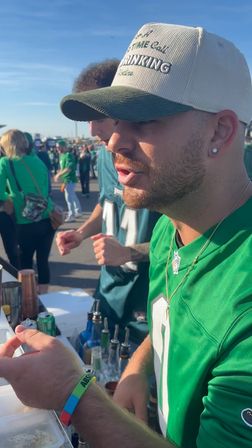 Close-up of a man in a green football jersey and baseball cap mixing drinks at a sunny tailgate in a crowded stadium parking lot, fans in green visible in the background.