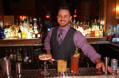 Smiling bartender in a vest and tie behind an illuminated, liquor-lined bar counter with three craft cocktails — a frothy coupe, a citrus rocks glass, and a tall rosemary-garnished highball in a cozy nightlife cocktail bar scene.