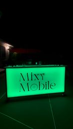 Glowing green illuminated portable bar sign on a dark outdoor patio at night, featuring sleek modern lettering and a small wheel graphic.