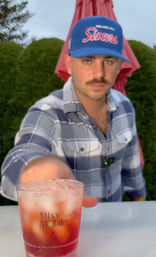 Person in a blue "Sixers" baseball cap and plaid shirt reaches a red iced cocktail in a clear plastic cup across an outdoor patio table with a red umbrella and green hedges behind.
