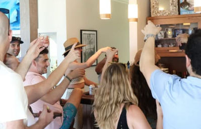 Cheerful group of friends in summer outfits raising drinks for a toast at a casual indoor bar or vacation rental.