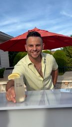 Smiling man offering a lemon-garnished cocktail at a backyard patio bar under a red umbrella, sunny summer outdoor gathering vibe