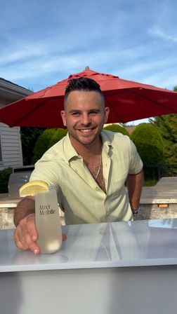 Smiling man offering a lemon-garnished cocktail at a backyard patio bar under a red umbrella, sunny summer outdoor gathering vibe