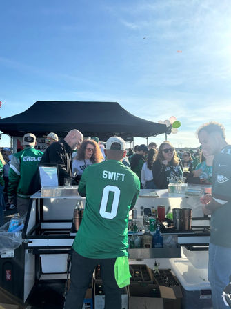 Outdoor Eagles tailgate in a stadium parking lot: fans in green jerseys crowd a drink cart under a black canopy, a person wearing a 'Swift 0' jersey serves beverages in late-afternoon sun.