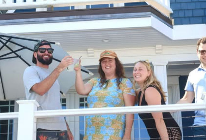 Four friends on a sunny beach house balcony cheering with drinks, smiling in casual summer clothes under a patio umbrella