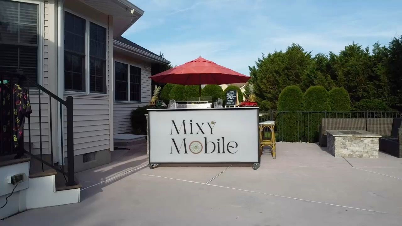 Wheeled outdoor bar with a red umbrella set up on a sunny suburban backyard patio, with stools, a stone firepit and tall hedges in the background.