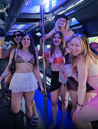Five women in swimsuits enjoying cocktails on a neon-lit party bus around a chrome dance pole — girls' night/bachelorette vibe