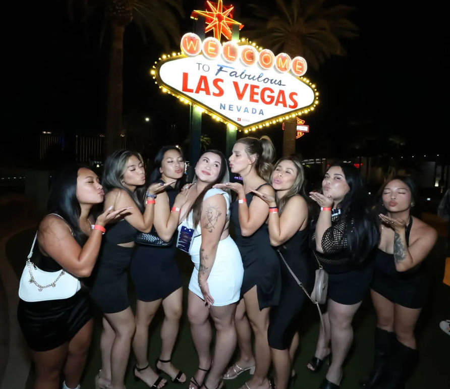 Group of women blowing kisses while posing in front of the glowing Welcome to Las Vegas, Nevada sign at night, lively nightlife photo.