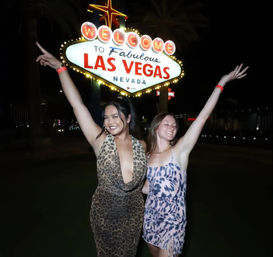 Two friends in patterned dresses smiling with arms raised at night in front of the iconic Welcome to Fabulous Las Vegas Nevada sign
