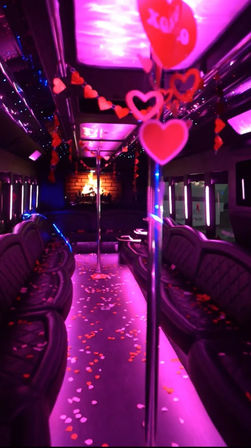 Valentine-themed party bus interior with glowing pink and purple LED lights, leather lounge seating, chrome dance poles, hanging heart garlands and heart-shaped confetti scattered down the aisle.