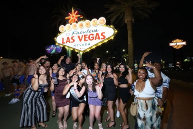 Group of friends posing and celebrating at night in front of the illuminated Welcome to Fabulous Las Vegas Nevada sign on the Las Vegas Strip, with palm trees and city lights in the background.
