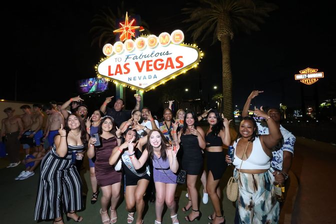 Group of friends posing and celebrating at night in front of the illuminated Welcome to Fabulous Las Vegas Nevada sign on the Las Vegas Strip, with palm trees and city lights in the background.