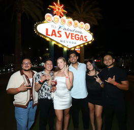 Six young adults posing at night beneath the neon 'Welcome to Fabulous Las Vegas Nevada' sign, smiling and holding drinks.