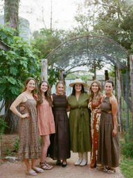 Six women in boho summer dresses smiling and posing arm-in-arm under a rustic metal arbor on a leafy garden path, one wearing a wide-brim hat and white boots.