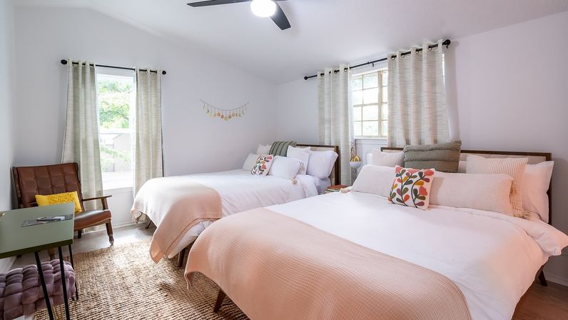Sunlit modern guest bedroom with two pale-pink layered beds, floral throw pillows, mid-century leather chair, woven rug, sheer curtains and black ceiling fan