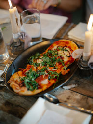 Candlelit flatbread pizza with melted mozzarella, pepperoni, fresh arugula and balsamic drizzle on a black platter over a rustic wooden table in a cozy restaurant setting