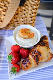 Picnic-style breakfast plate: shrimp-topped creamy grits in a bowl, chocolate-chip croissant and fresh strawberries beside a wicker picnic basket on a striped tablecloth