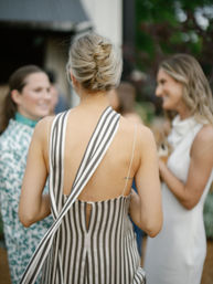 Back view of a woman in a backless black-and-white striped dress with a twisted updo, chatting with friends at an outdoor garden party