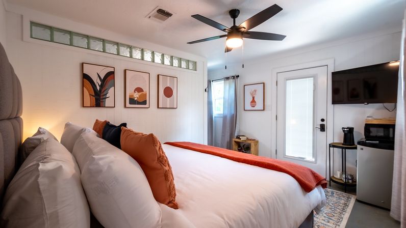 Sunlit cozy modern bedroom with large bed, terracotta throw and pillows, abstract wall art, ceiling fan, wall-mounted TV and mini-fridge with coffee station.